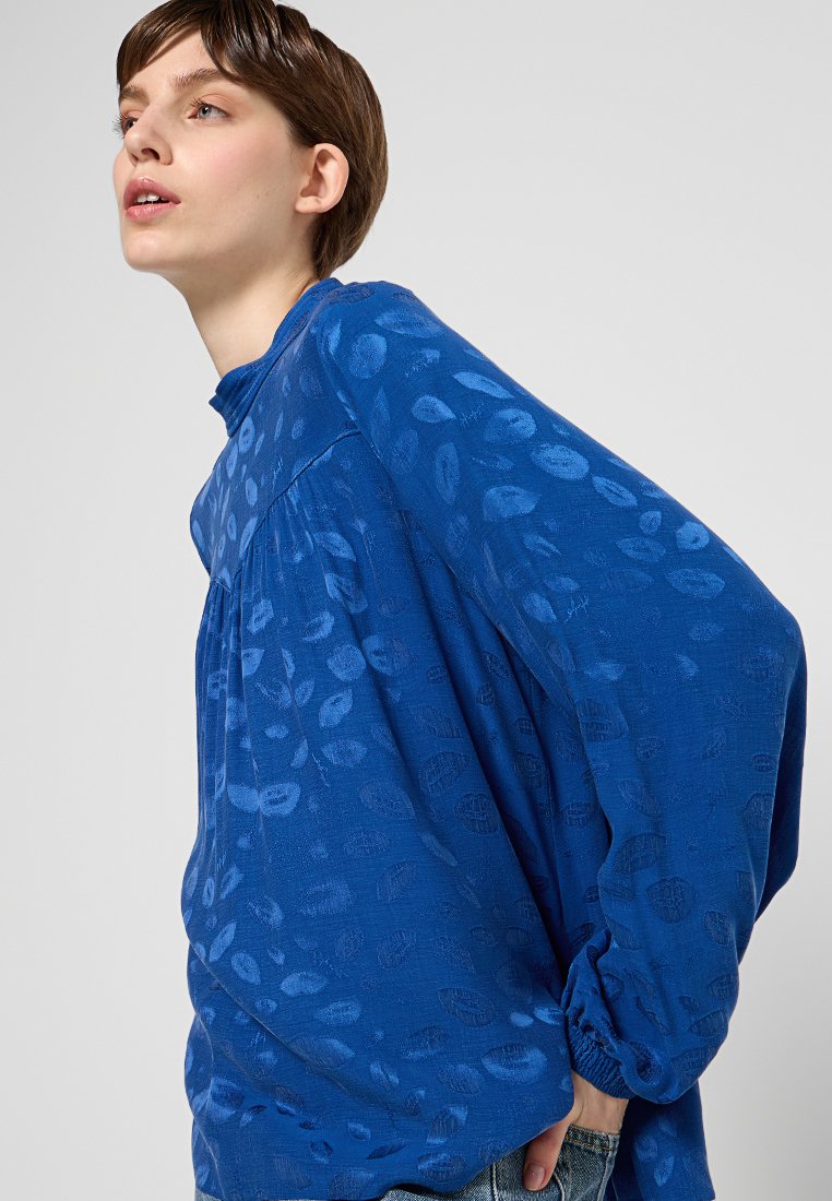 Blaues Langarmoberteil aus leichtem Stoff, mit einem strukturierten Muster aus abstrakten Formen, gerafftem Schulterdetail und geknöpften Ärmeln.