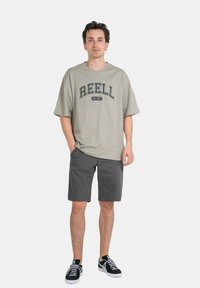 Gray oversized t-shirt with "REELL" text graphic, paired with dark gray shorts and black sneakers. Simple design, casual fit, cotton material.