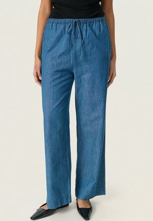 Mujer con pantalones vaqueros azules de pierna ancha, cintura elástica y cordón, combinados con zapatos planos negros de punta puntiaguda y un top negro.