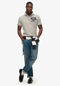 Grijze poloshirt met marineblauwe tekst, blauwe jeans en zwarte schoenen. Heeft een stijlvolle kraag en opgerolde mouwen, gelaagd met een marineblauw en witte trui.