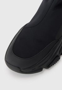 Chaussure de sport noire avec une tige textile lisse, une semelle en caoutchouc et un design rembourré, avec des coutures contrastées au niveau des orteils.