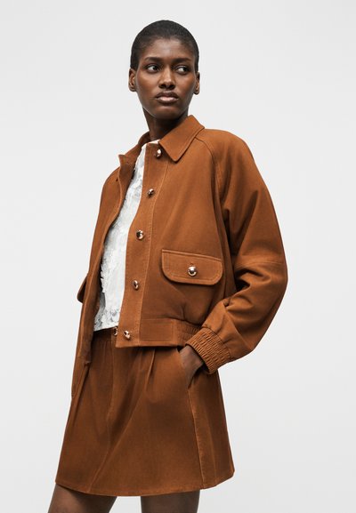 Γυναίκα που φοράει σακάκι σε χρώμα σκουριάς με κουμπιά και τσέπες με καπάκι, καθώς και φούστα στο ίδιο χρώμα, ποζάροντας με το ένα χέρι στην τσέπη, με λευκή δαντελένια μπλούζα από κάτω.