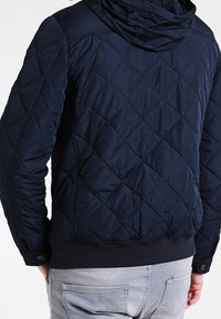 Mörkblå quiltad jacka med huva, diagonala sömmar, elastiska ärmslut och ribbat nederkant för en snug passform. Lämplig för kyligt väder.