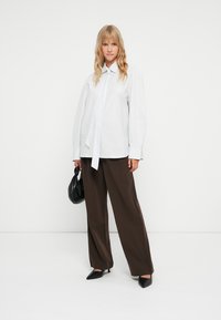 Camicia bianca oversize con dettaglio cravatta, abbinata a pantaloni ampi marrone scuro. Ballerine nere con punta e una piccola borsa nera completano il look.