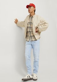 Beige bomberjack over een geruite blouse, lichtblauwe jeans, rode pet en witte sneakers met zwarte strepen; ontspannen fit, casual outfit.
