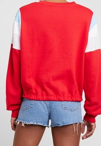 Červená mikina s lehkými modrými a bílými pruhy na rukávech, s žebrovaným výstřihem a stahovaným lemem. Doplněná o džínové šortky.