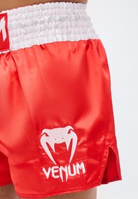 Κοντινή λήψη κόκκινων σατέν μποξερό με λευκή ελαστική μέση και λευκό λογότυπο Venum στο αριστερό πόδι.