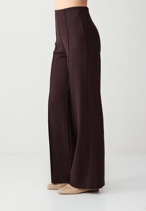 Spodnie o wysokim stanie i szerokich nogawkach w ciemnym brązie, noszone z beżowymi płaskimi butami, pokazane z boku na jasnym, jednolitym tle.