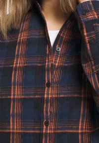 Camicia a quadri blu e arancione con design a bottoni, taschino sul petto e bottoni neri realizzata in un morbido tessuto testurizzato.