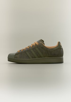 Baskets basses Adidas vert olive avec une texture crocodile embossée, lacets beiges et logo Adidas sur la languette du talon.