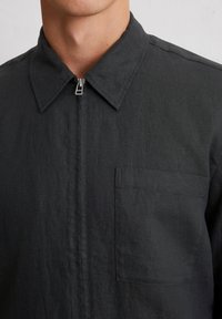 Black zip-up shirt with a structured collar, single chest pocket, and a textured fabric. Minimalist design and clean lines.