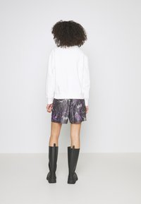 Svarta gummistövlar i knähöjd, vit sweatshirt och shorts med lila blixtmönster. Modell med ryggen mot kameran; lockigt hår synligt. Enkelt bakgrund.