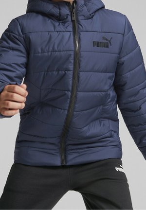 Marineblå pufferjakke med lynlåsåbning, quiltet struktur og hætte. Har sort Puma-logo på brystet og sort lynlås.