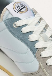 Zapatilla azul clara con detalles de ante en gris y azul, cordones blancos y un parche redondo con logo en la lengüeta. Materiales texturizados visibles.