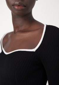 Närbild av en person med en svart ribbad topp med en vit kantad fyrkantig halsringning, som visar hals, axel och läppar.