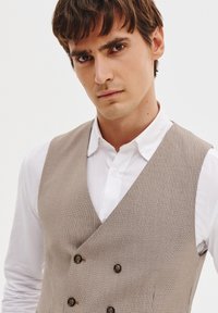 Beżowa kamizelka w wzór houndstooth z podwójnym zapięciem na guziki, noszona na białej koszuli z kołnierzykiem. Gładki materiał o dopasowanym kroju.