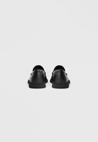 Zapatos negros de piel sin cordones con un acabado texturizado, punta redonda y suelas bajas vistos desde atrás. Diseño sencillo sin acentos visibles.