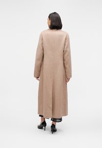 Μακρύ μπεζ παλτό από μάλλινο ύφασμα με ευθύγραμμη σιλουέτα, κεντρική ραφή και μακριά μανίκια. Φορεμένο πάνω από μαύρο φόρεμα με δαντελωτό τελείωμα και μαύρες γόβες.