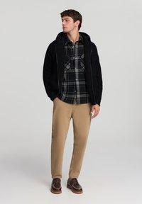 Czarna rozpinana bluza z teksturowanym kołnierzem, nałożona na ciemną koszulę w kratę, połączona z jasnobeżowymi spodniami i brązowymi butami.