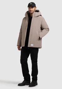 Vaatteena on vaahtoparka vaaleanbeigellä värillä, hupulla ja etutaskuilla, yhdistettynä mustiin housuihin ja kenkiin. Yksinkertainen design, jossa on vain vähän korostuksia.