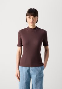 Braunes, geripptes Kurzarmoberteil mit hohem Kragen und subtilen Markenakzenten. Glatte Textur, figurbetontes Design, kombiniert mit hellblauen, lockeren Jeans.