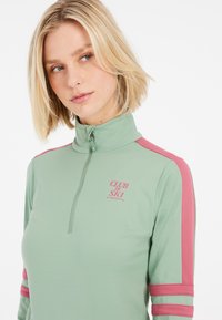 Svetlozelený zipsový top s ružovými pruhmi na rukávoch, so vzorom s nápisom "CLUB DE SKI." Hladká textúra a priliehavý strih.