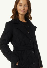 Black quilted coat with a shawl collar, belted waist, and two side pockets. Features large buttons and a textured fabric finish.
