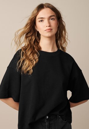 Mujer joven con el pelo largo y ondulado, que lleva grandes pendientes de aro y una camiseta negra holgada sobre un fondo beige liso.