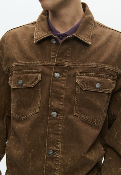 Καφέ τζιν μπουφάν με δύο τσέπες στο στήθος, ασημένια μεταλλικά κουμπιά, γιακά και φθαρμένη υφή. Το ύφασμα εμφανίζει διακριτικά μοτίβα φθοράς.