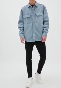 Camisa de denim azul claro con dos bolsillos en el pecho, botón frontal y mangas largas, combinada con pantalones negros y zapatillas blancas.