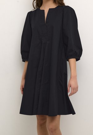 Vrouw die een losse zwarte jurk draagt tot op de knie met driekwartpofmouwen en een vierkante halslijn met een kleine voorklief.