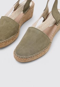 Sandalias de cuña de ante en verde oliva, con una suela de yute texturizada. Presenta correas elásticas trenzadas para un ajuste seguro y fácil de poner. Diseño de puntera redondeada.