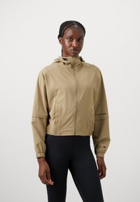 Kvinne med flette i håret, ikledd en beige glidelåshettejakke og svarte leggings, stående mot en enkel hvit bakgrunn.
