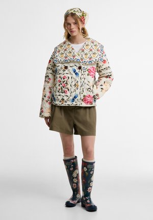 Nő virágos steppelt dzsekit, hozzáillő fejkendőt, olívazöld rövidnadrágot és tengerészkék virágos esőcsizmát visel, egyszínű háttér előtt áll.