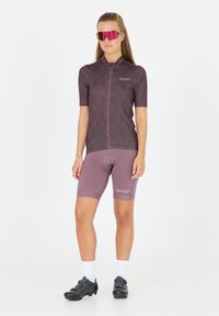 Tenue de cyclisme comprenant un maillot mauve à manches courtes et à fermeture éclair avec un motif à pois, accompagné de shorts assortis. Chaussures de cyclisme noires et lunettes de soleil teintées de rose.