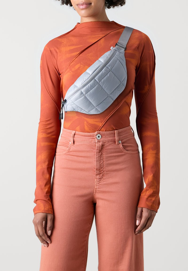 Stříbrná prošívaná crossbody taška se zipovým uzávěrem, nošená přes dlouhý rustový top s abstraktním vzorem, doplněná růžovými kalhotami.
