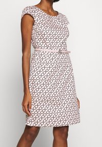 Robe rose avec un motif circulaire noir répétitif, des manches courtes, une coupe ajustée et une ceinture fine assortie avec une boucle ronde.