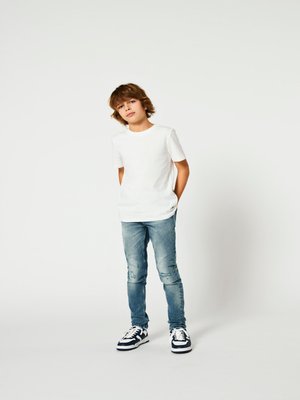 Maglietta bianca a maniche corte, jeans strappati blu chiaro e sneakers bianche e nere. Modello in piedi con le mani in tasca su uno sfondo neutro.