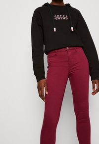 Zwarte cropped hoodie met roze logo en trekkoorden, gecombineerd met bordeauxrode skinny jeans. Gladde textuur, minimalistisch ontwerp.