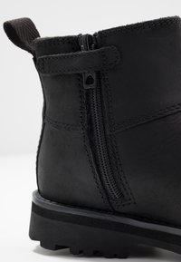 Botín de cuero negro hasta el tobillo con un cierre lateral, costura reforzada, superficie texturizada y una suela de goma duradera que presenta un patrón de relieve.