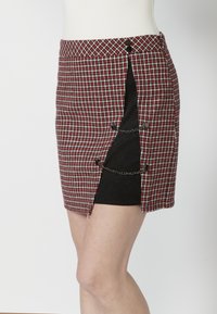 Mini-jupe à carreaux rouge et noir avec fente latérale, ornée de boutons en métal et de détails en chaîne, fabriquée en tissu texturé.