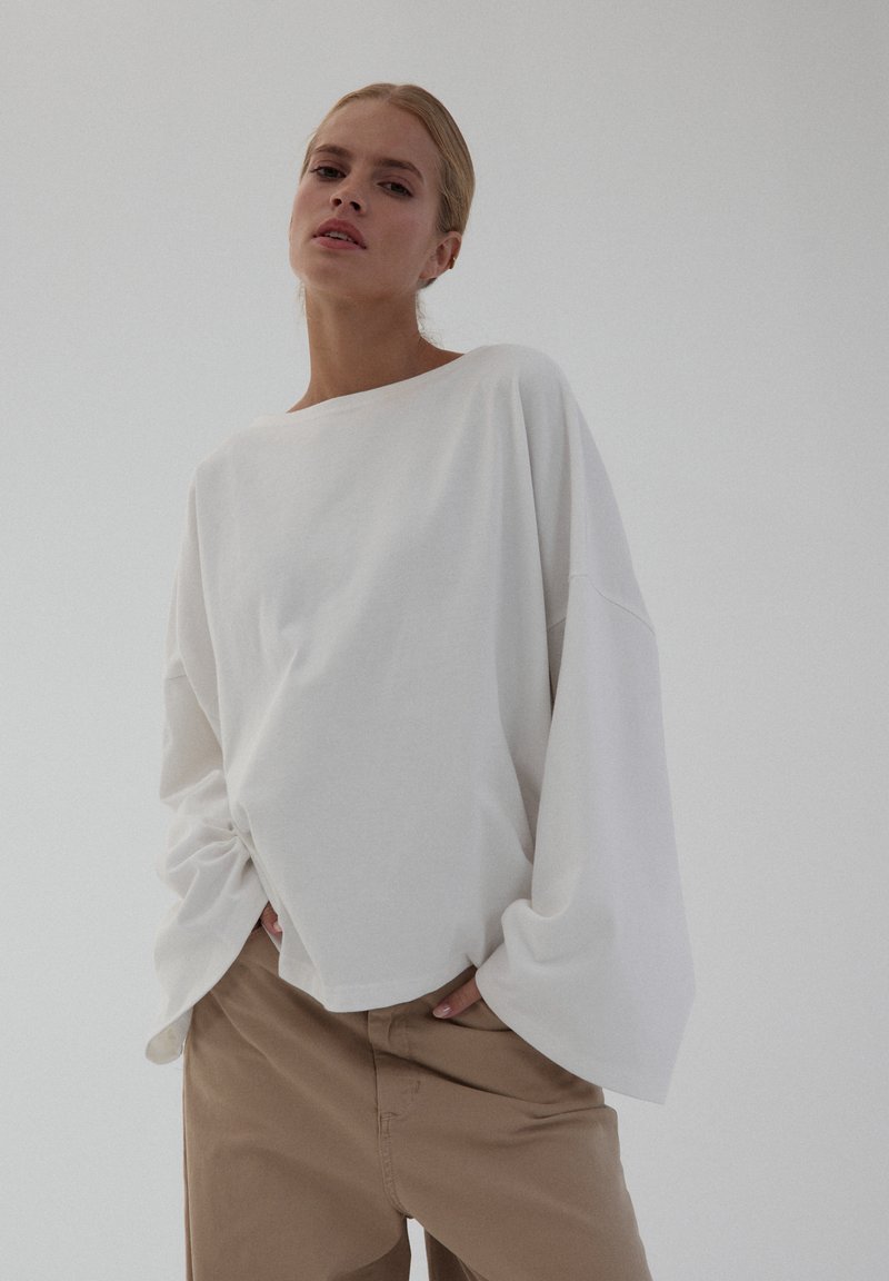 Biała, oversize'owa bluzka z długim rękawem, okrągłym dekoltem i opuszczonymi ramionami, w połączeniu z beżowymi spodniami. Gładka faktura, minimalistyczny design.