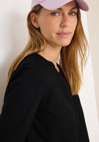 Schwarze Bluse mit rundem Ausschnitt und langen Ärmeln. Das Model trägt eine hellrosafarbene Kappe. Der Stoff wirkt glatt mit einer weichen Textur.