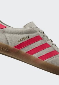 Gray suede sneaker with pink stripes, gum sole, "GAZELLE" text, and lace-up design.