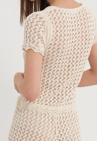 Kremowy sweter dziany z koronkowym wzorem, krótkimi rękawkami z falistymi brzegami oraz dopasowanym krojem podkreślającym fakturę i kształt.
