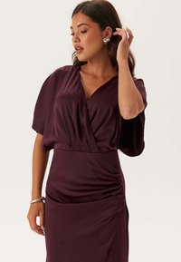 Kvinna i en djupt lila omslagsklänning med rynkad detalj på sidan, bärande silvriga hjärtformade örhängen och ett silverarmband, poserande med handen nära örat.