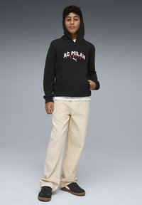 Sweat à capuche noir avec "AC MILAN" et logo Puma en blanc/rouge, poche avant, associé à un pantalon couleur crème et des baskets noires sur un fond dégradé.