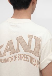 Person von hinten gesehen, trägt ein helles T-Shirt mit großen strukturierten Buchstaben "CANU" und kleinerem Text "Originator of streetwear".