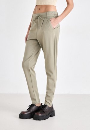 Pantalon beige clair avec taille à cordon de serrage et poches avant, associé à des chaussures montantes en cuir brun foncé à lacets. Texture de tissu lisse.