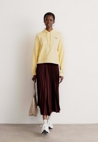 Felpa gialla con cappuccio, gonna midi plissettata borgogna, sneakers bianche e borsa beige. Presenta un tessuto testurizzato e un design dall'aspetto rilassato.